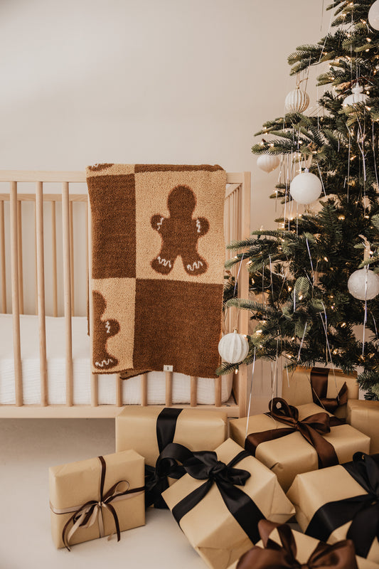 A cozy nursery scene features a light wood crib draped with the Gingerbread Checkerboard Plush Blanket by forever french baby. Nearby, an OEKO Standard 100 certified Christmas tree is adorned with white ornaments and ribbons. Several wrapped gifts with dark bows rest underneath.