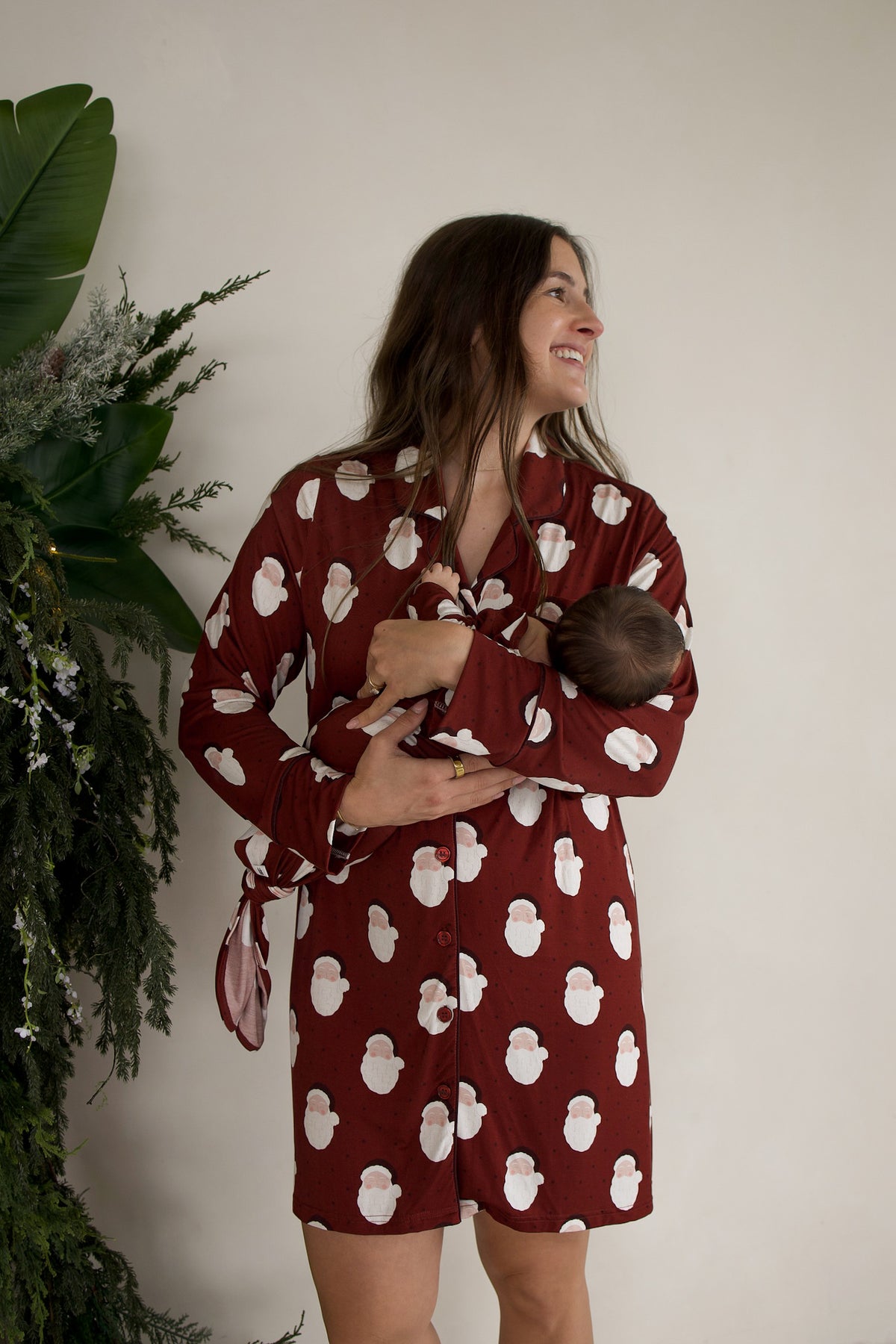 A woman wearing a matching maroon button-down shirt with a Santa Claus pattern holds a newborn baby in a Bamboo Knotted Gown | Vintage Santa.