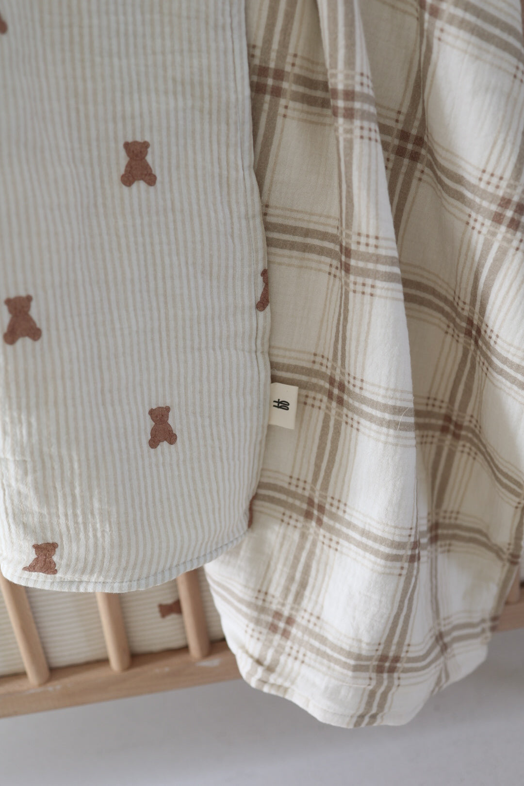 Close-up of nursery bedding in a crib featuring the forever french baby Muslin Quilt | Teddy & Ellis Plaid: one side displays small brown teddy bears on stripes, while the other has a cozy brown and beige plaid pattern.