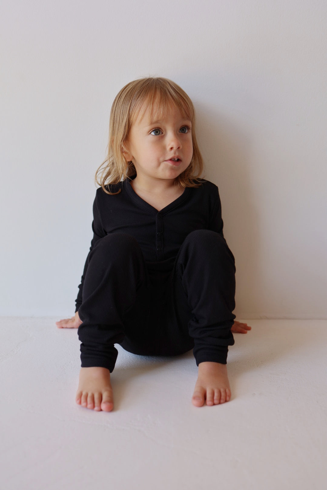 A young child with shoulder-length blonde hair, dressed in Forever French Baby's Bamboo Two-Piece Pajamas in Black Ribbed, is sitting against a white wall. The child's legs are bent, feet flat on the ground, and hands resting between their legs. The child gazes into the distance with a thoughtful expression.