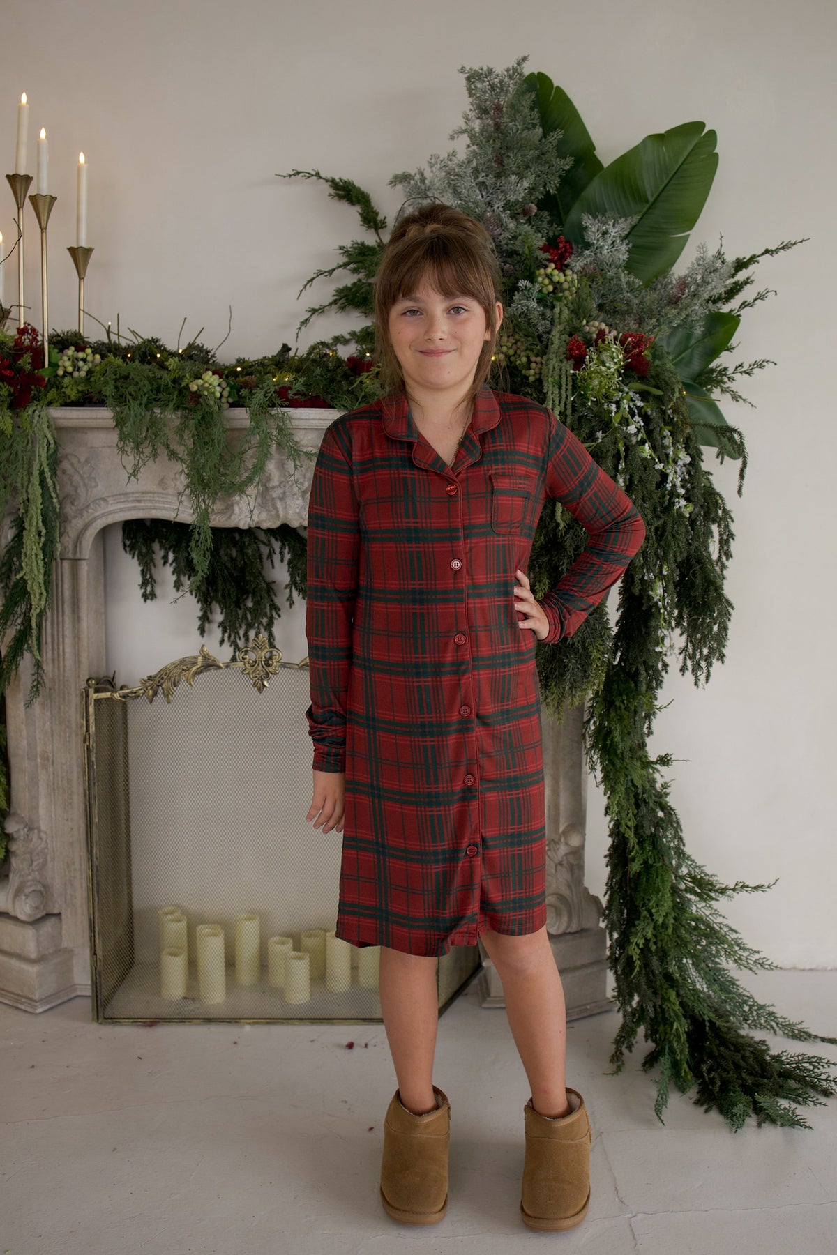 A child wearing a Bamboo Sleeping Dress | Christmas Plaid and tan ankle boots, standing in front of a fireplace decorated for the holidays.
