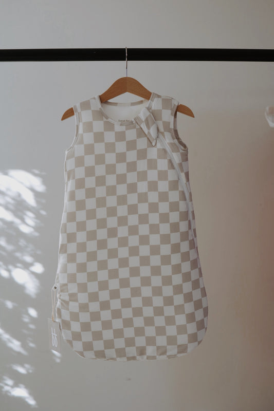 The Bamboo Sleep Sack | Checkerboard by forever french baby, in beige and white checks, hangs on a wooden hanger against a plain background with soft sunlight casting shadows on the left.