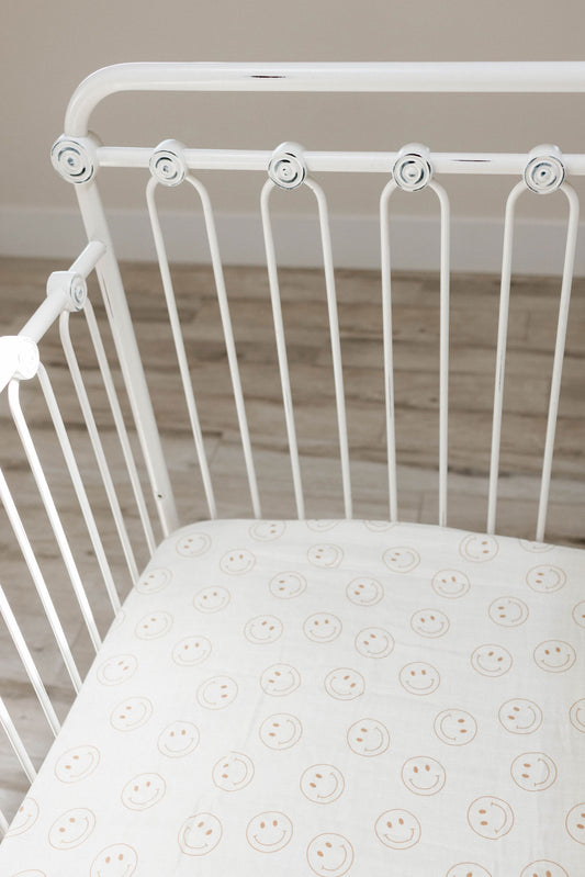 A white metal crib with a crib mattress covered in Forever French Baby's Muslin Crib Sheet, featuring the Just Smile Ivory design in 100% muslin cotton. The crib is set on a wooden floor with a light finish, creating a minimalistic and calm overall scene.