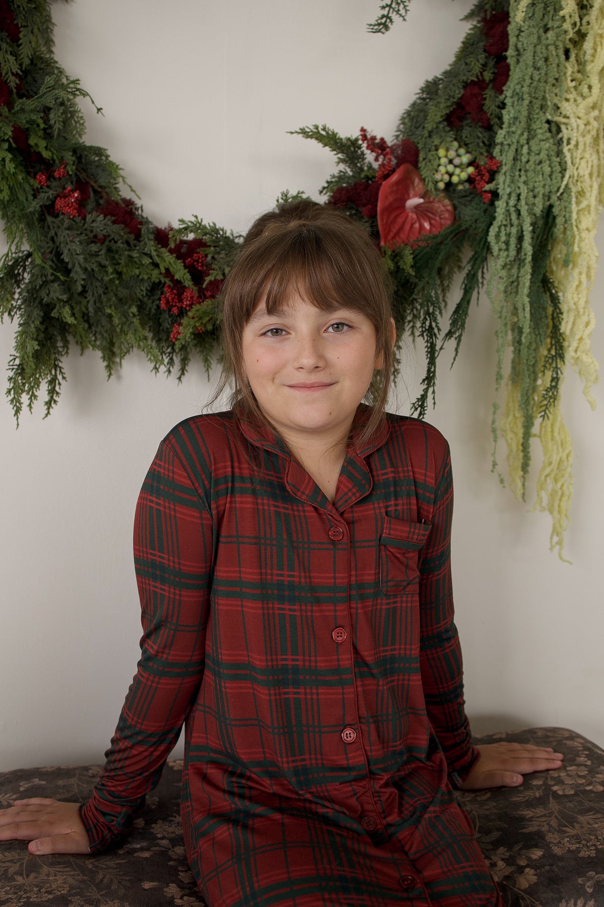 A young girl smiles in front of a festive wreath, wearing the Bamboo Sleeping Dress | Christmas Plaid by forever french baby—soft, hypoallergenic children’s clothing against a plain white wall.