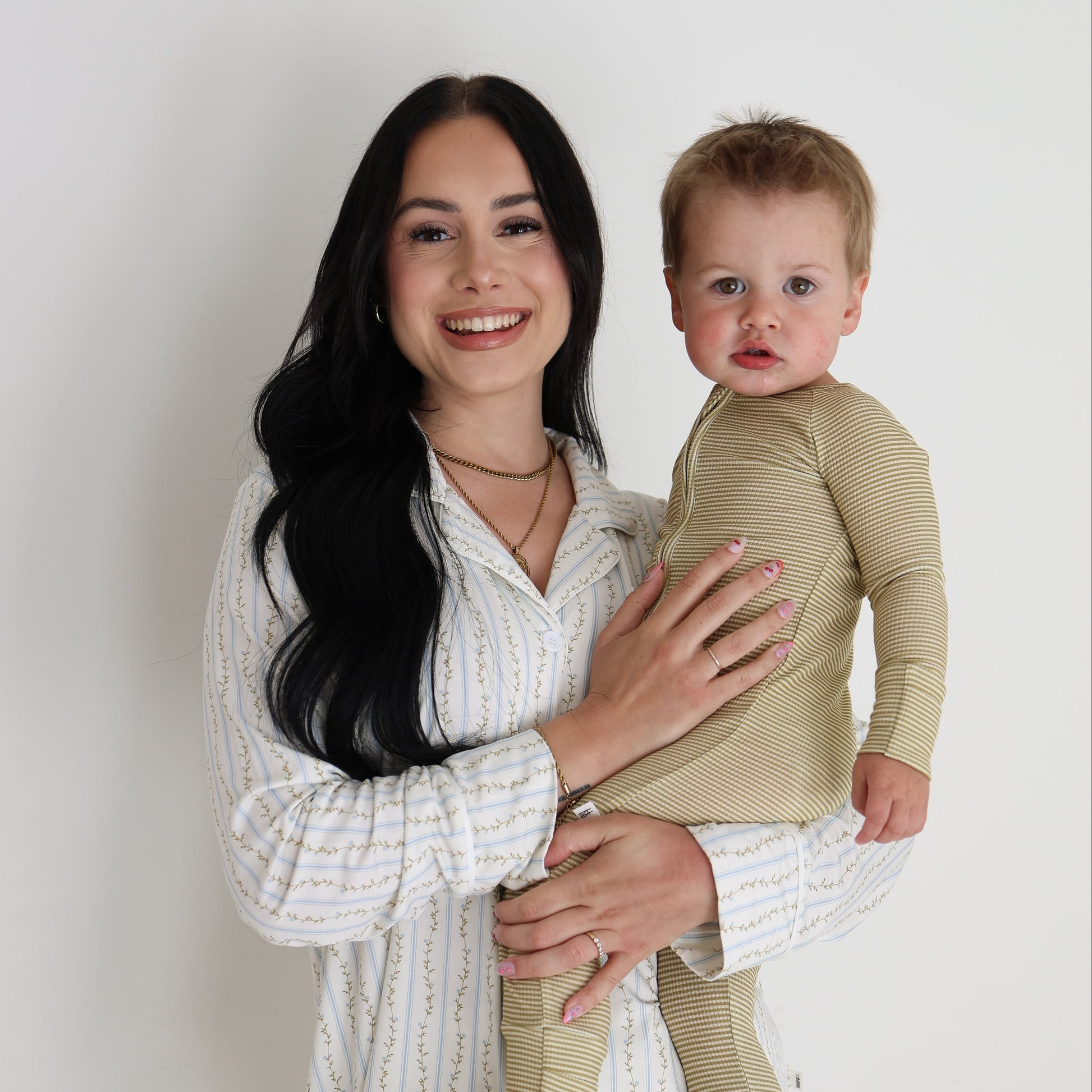 A smiling woman in a white patterned shirt holds a young child dressed in forever french baby’s Bamboo Zip Pajamas | Willow Stripe Ribbed, standing against a plain white background.