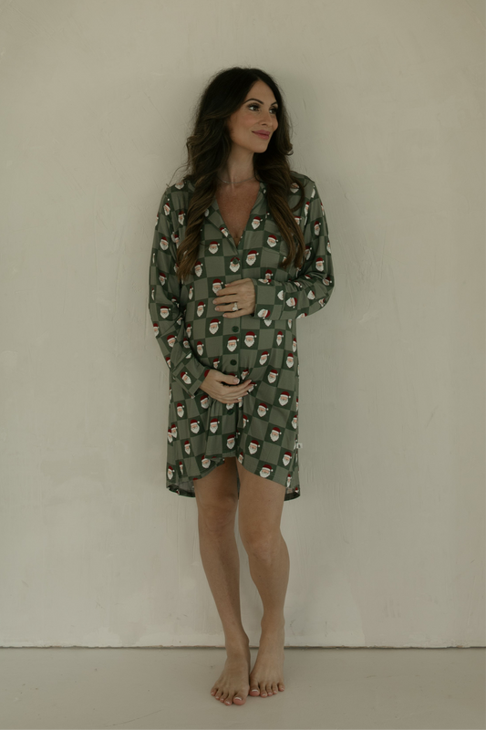 A pregnant woman with long brown hair stands barefoot against a light wall, smiling softly as she cradles her belly. She wears the forever french baby Women's Bamboo Sleeping Dress in the Santa Checkerboard pattern.