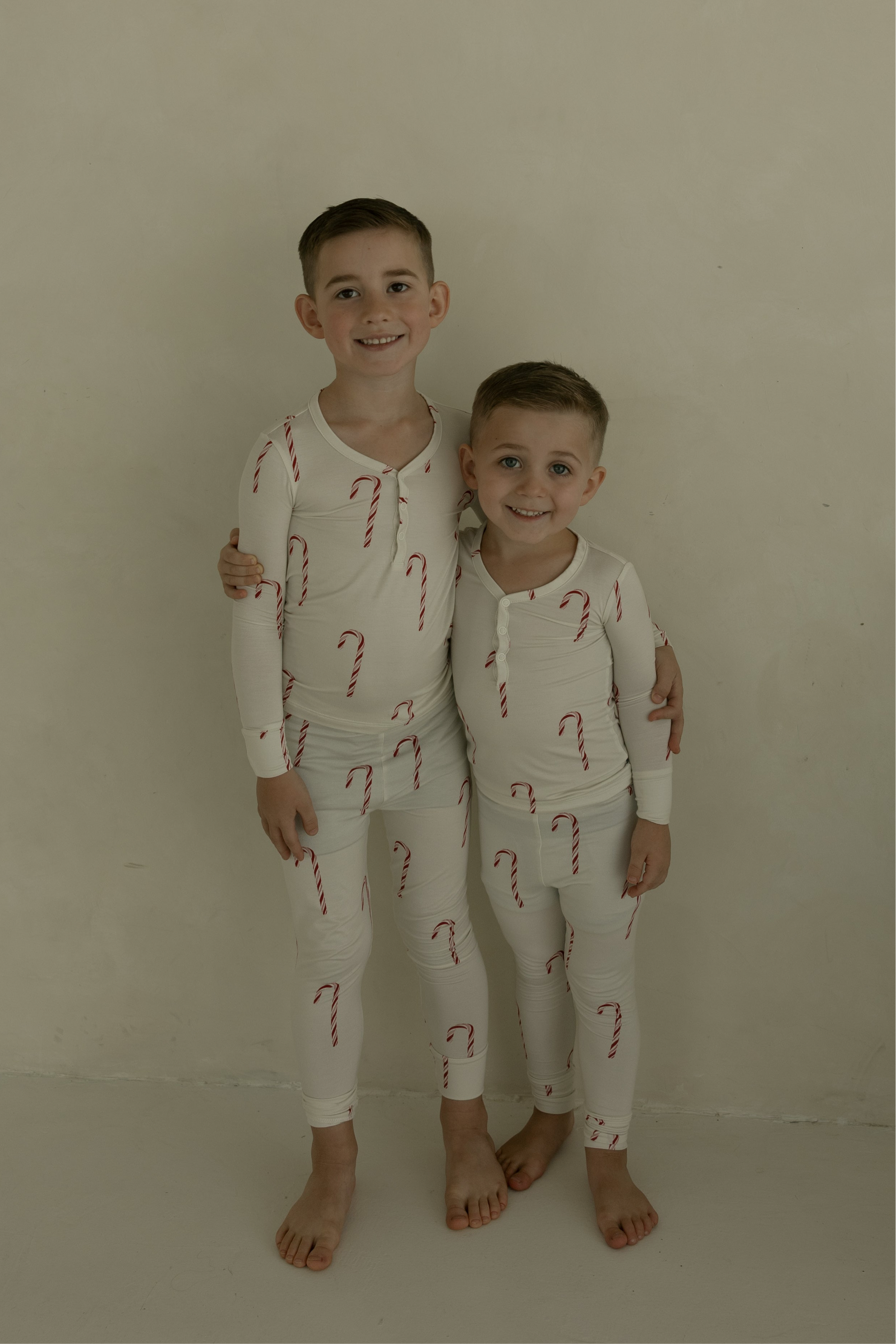 Two young boys smile and hug barefoot against a light wall, wearing matching forever french baby Bamboo Two Piece Pajamas | Candy Cane—a cozy, festive choice for kids’ sleepwear.