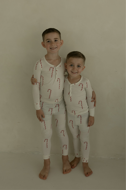 Two young boys smile and hug barefoot against a light wall, wearing matching forever french baby Bamboo Two Piece Pajamas | Candy Cane—a cozy, festive choice for kids’ sleepwear.