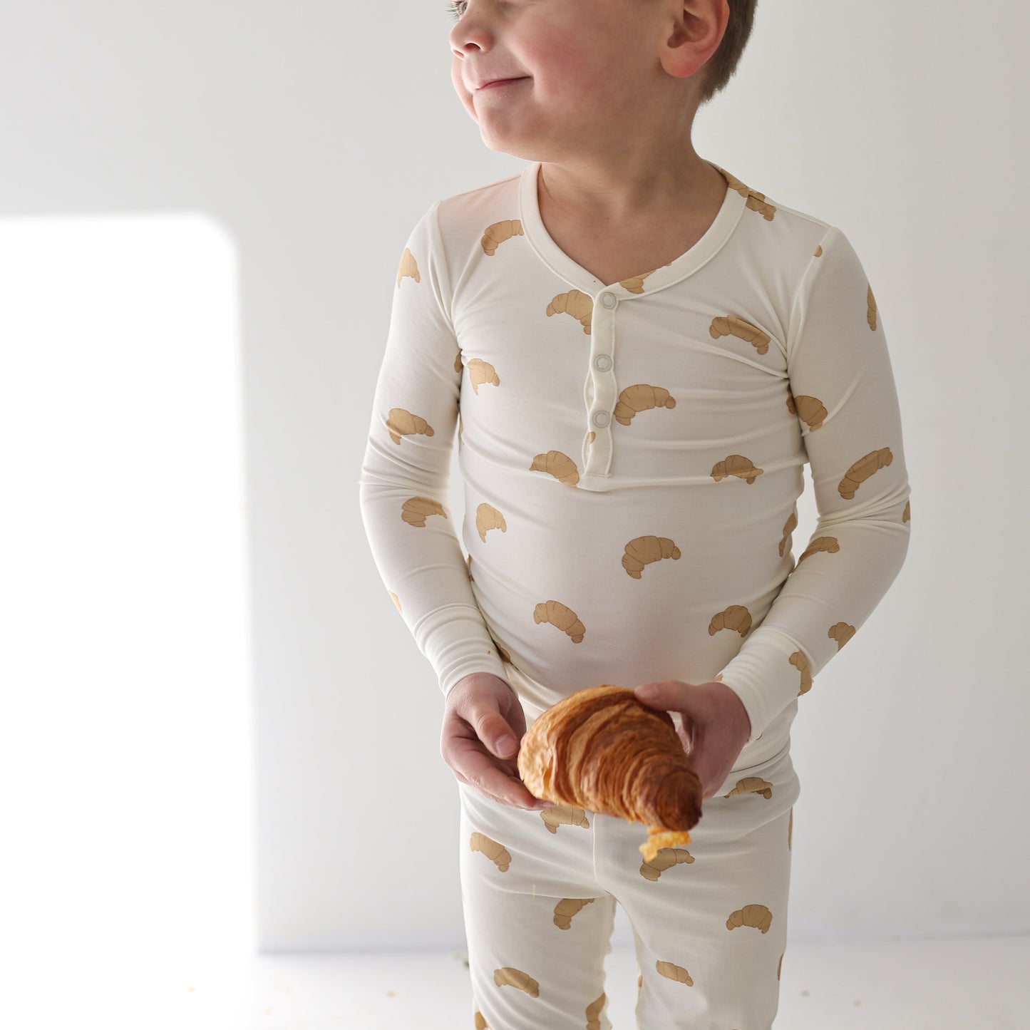 A smiling child in forever french baby’s Bamboo Two Piece Pajamas | Croissants, featuring a playful croissant print, holds a large croissant. The breathable sleepwear pops against a bright, minimalistic background.