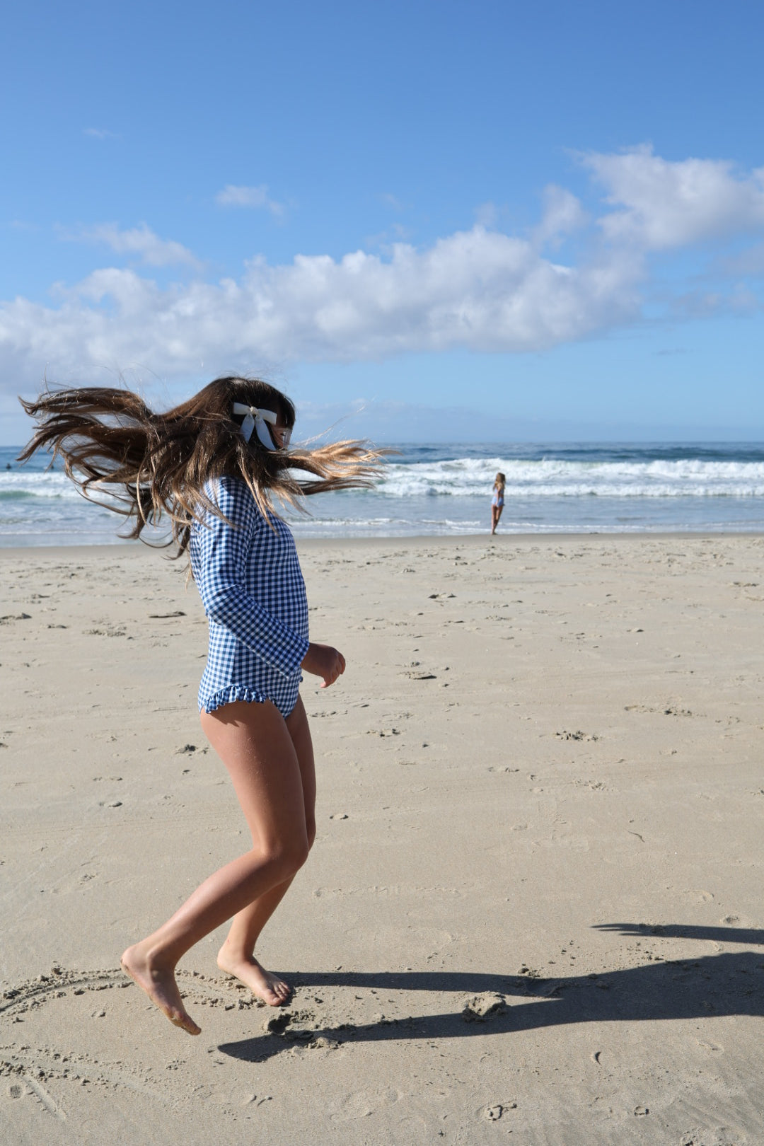 A girl wearing the forever french baby Girls Long Sleeve Swimsuit in Blue Gingham and a white hair bow spins on the sandy beach, with another person by the shoreline and waves under a partly cloudy sky.