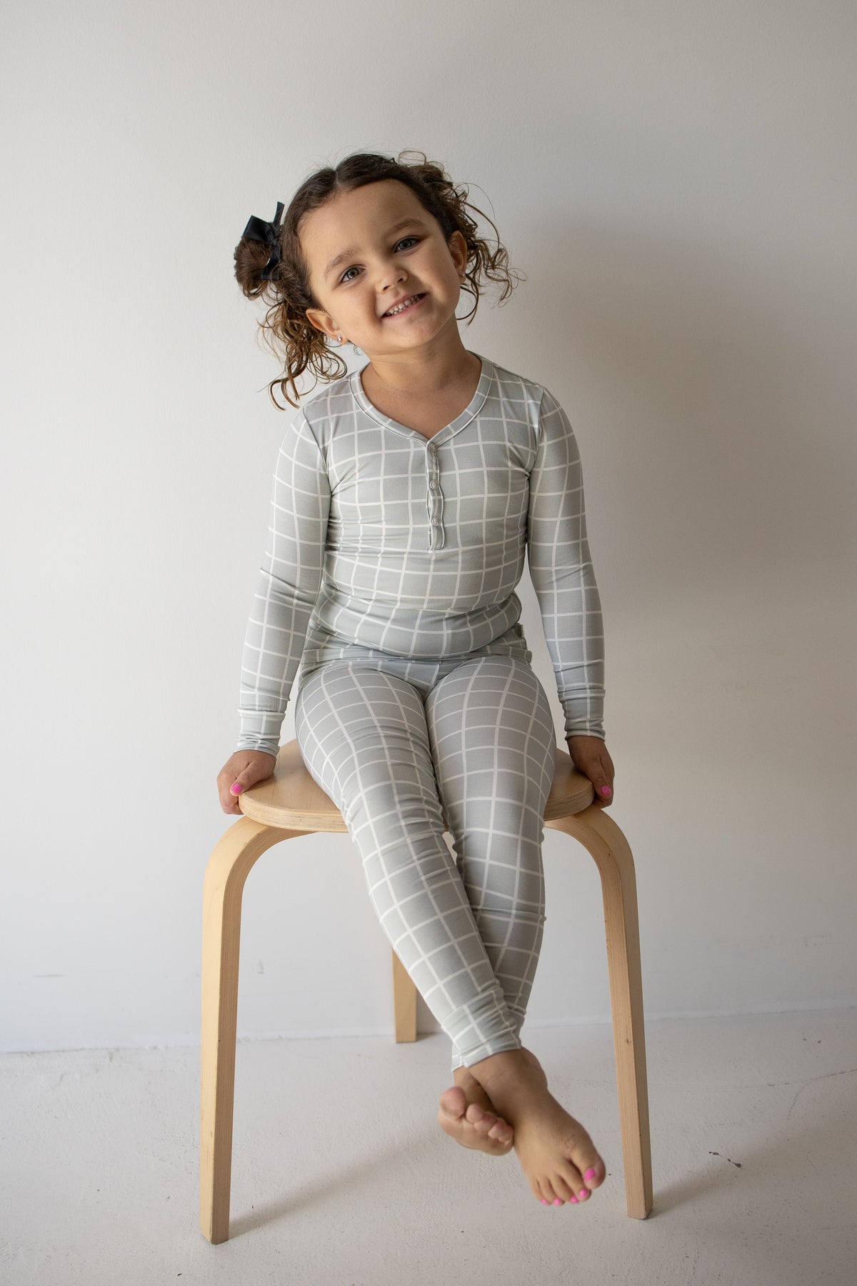 A young child with curly hair, dressed in the forever french baby Bamboo Two Piece Pajamas in the Foggy Grid pattern, sits barefoot on a wooden stool against a plain white wall, smiling at the camera.