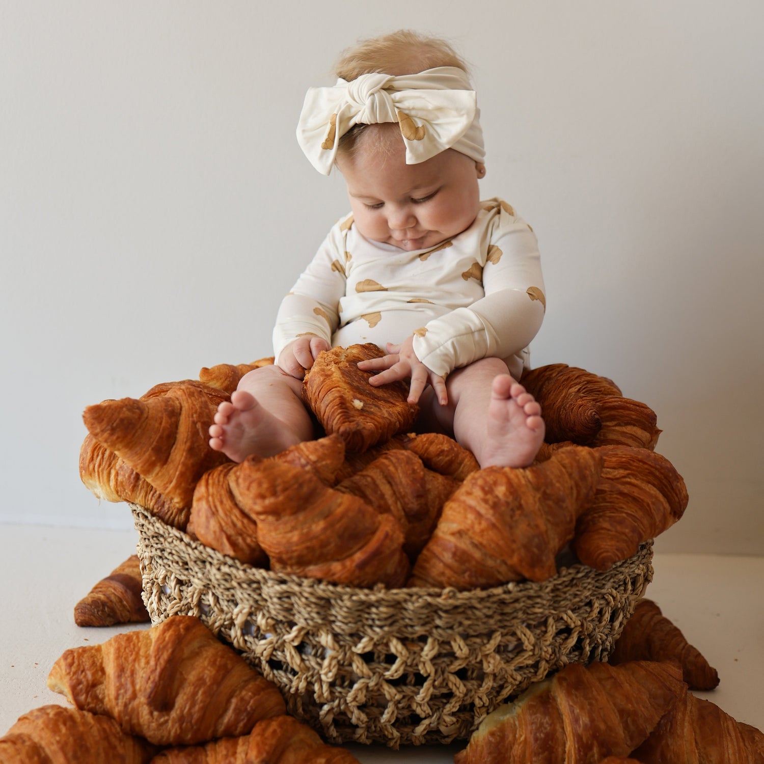 A baby in a white headband and the forever french baby Long Sleeve Bamboo Snapsuit | Croissants sits in a woven basket filled with croissants, gazing at the pastries with feet resting on them while extra croissants are scattered nearby.