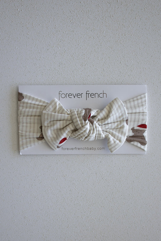 A cream and beige plaid Bamboo Head Wrap in the Santa Teddy style, featuring a large bow, is displayed on a "forever french baby" branded card against a light textured wall.
