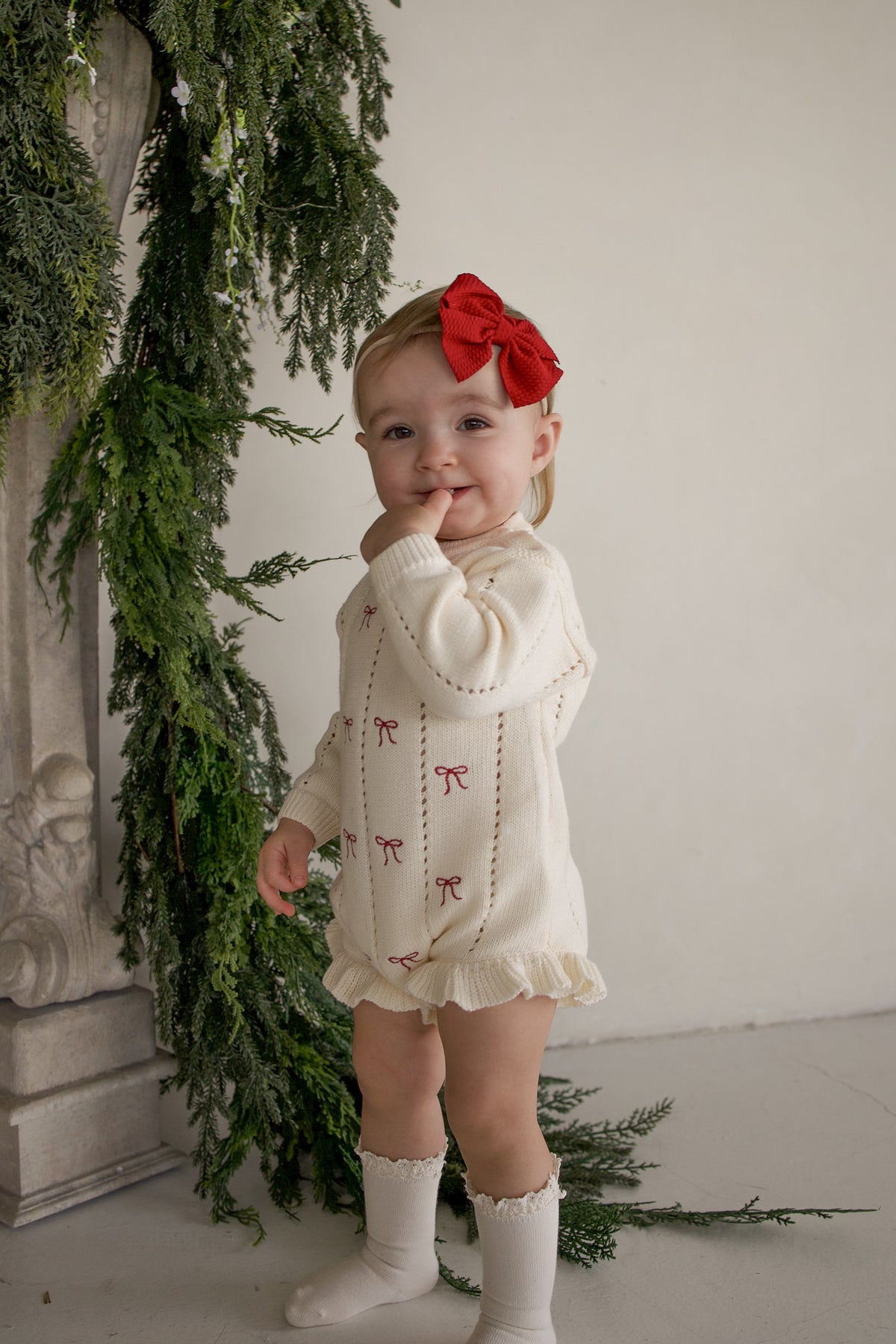 A toddler wears a cream-colored Knit Ruffle Romper | Candy Cane with red bow patterns, a matching red hair bow, and ruffled socks.