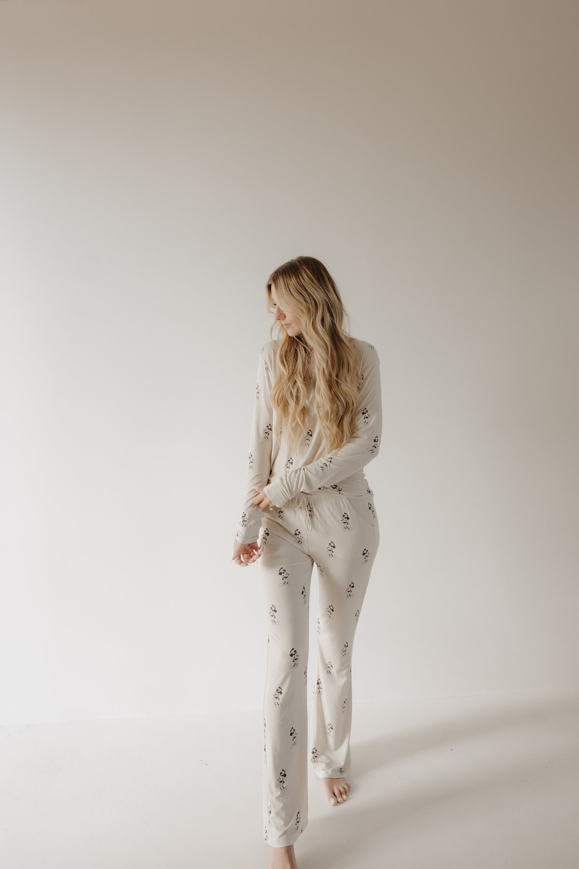 A woman with long, wavy blonde hair stands barefoot against a white background, wearing forever french baby Women’s Bamboo Pajama | All Aboard!, which features a light color and subtle dark pattern. She looks left, arms relaxed.
