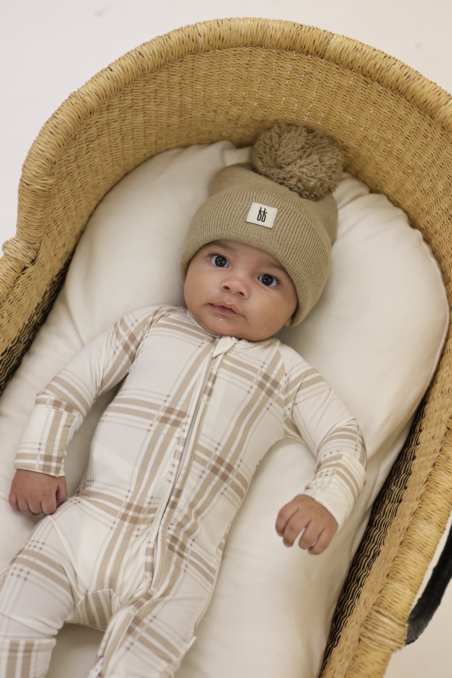 A baby in forever french baby's Bamboo Zip Pajamas | Ellis Plaid and a tan pom-pom beanie lies in a wicker bassinet with a white cushion, looking up at the camera.