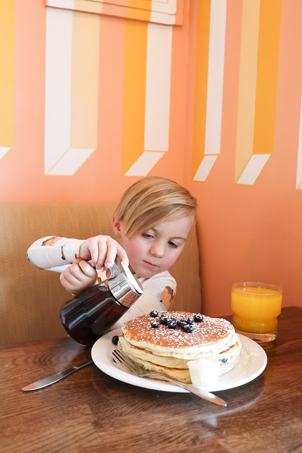 A young child in forever french baby Bamboo Two Piece Pajamas | Sunday Stack pours syrup onto blueberry pancakes with powdered sugar, a glass of orange juice by their side, seated at a wooden table in a vibrant, striped-walled cafe.