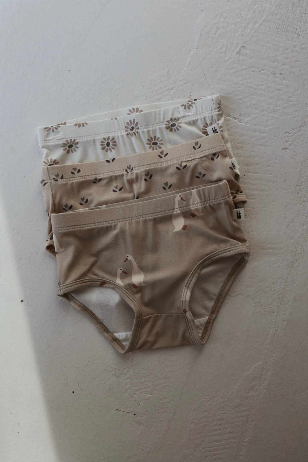 Three pairs of forever french baby Girl's Bamboo Briefs (Flora, Silly Goose & Daisy Fields) in soft neutral tones, stacked on a textured surface with gentle natural light. Hypo-allergenic and perfect for girls.