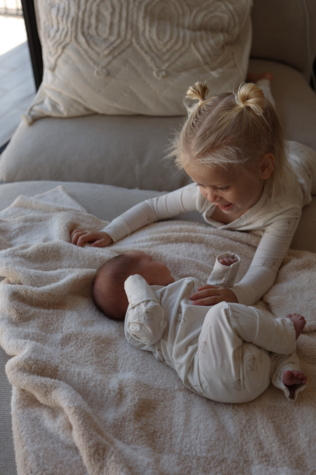 A young blonde girl in pigtails smiles at a baby on a beige blanket. Both kids wear forever french baby Bamboo Two Piece Pajamas | Dandelion while relaxing on a cozy sofa with a textured pillow in the background.