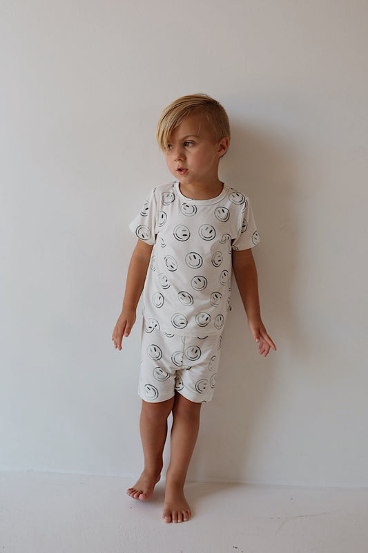 A young child stands barefoot against a white wall, wearing the forever french baby Bamboo Short Set in Green Smiles—breathable sleepwear with black smiley face prints—looking to the side with arms slightly outstretched.