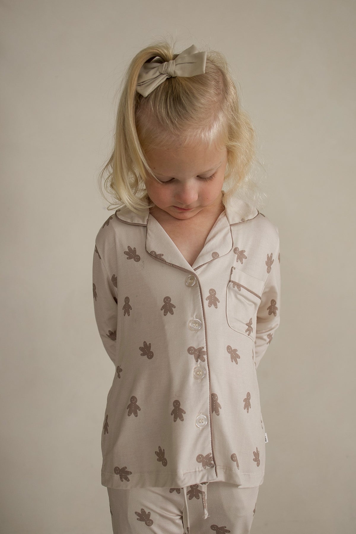 A child wearing a Girl’s Bamboo Button Up Lounge Set | Gingersnap, featuring a light-colored, long-sleeve pajama set with a gingerbread man print.
