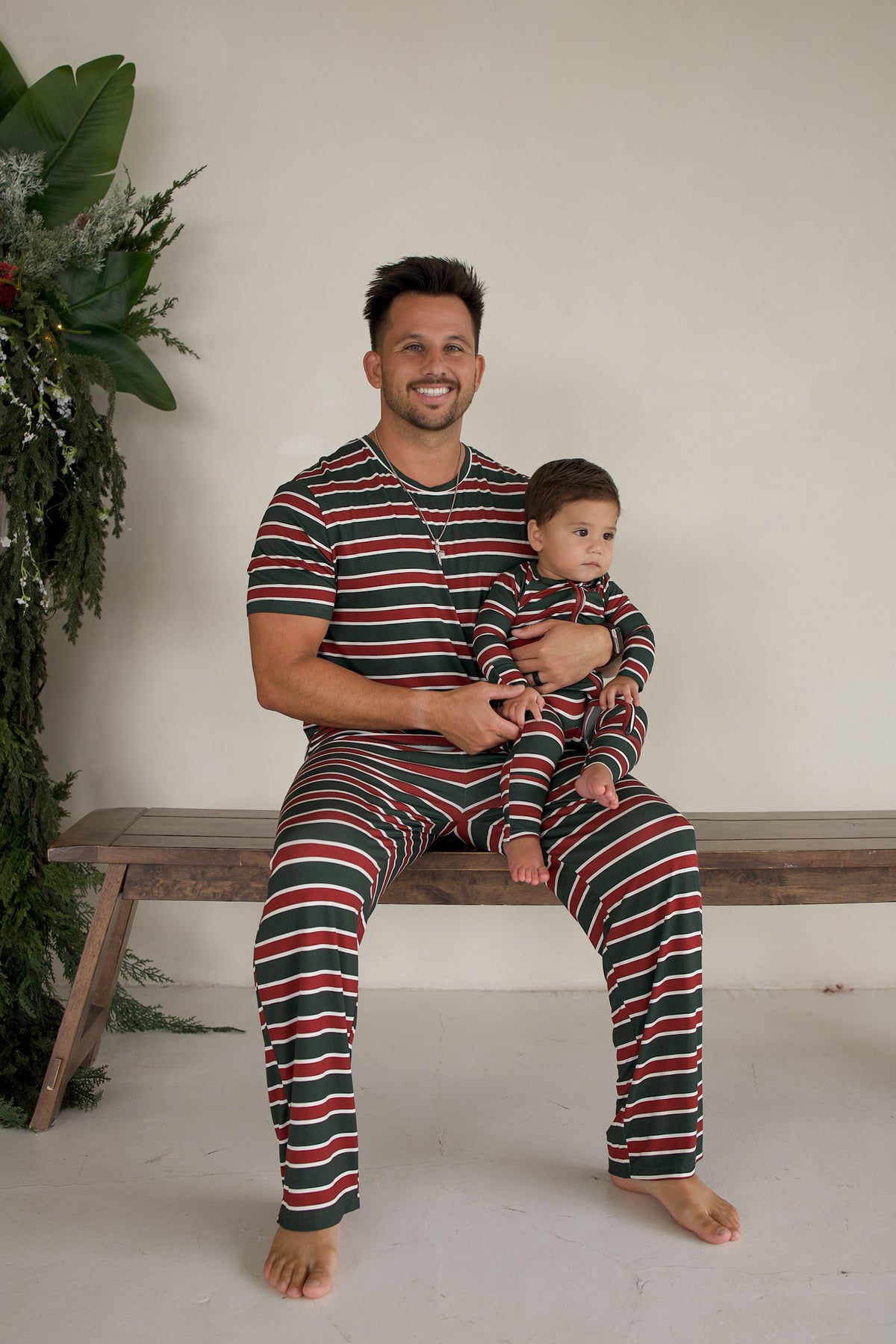 A man and an infant sit on a bench wearing matching Men’s Short Sleeve Pajamas | Holiday Stripe in a red, green, and white striped pattern.