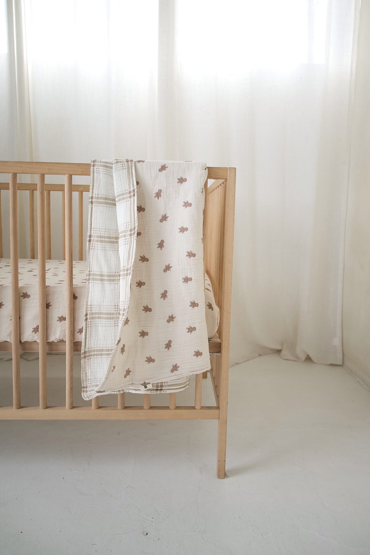 A reversible forever french baby Muslin Quilt | Gingersnap & Ellis Plaid with a gingerbread man print on one side and a neutral plaid pattern on the other, draped over the side of a wooden crib.