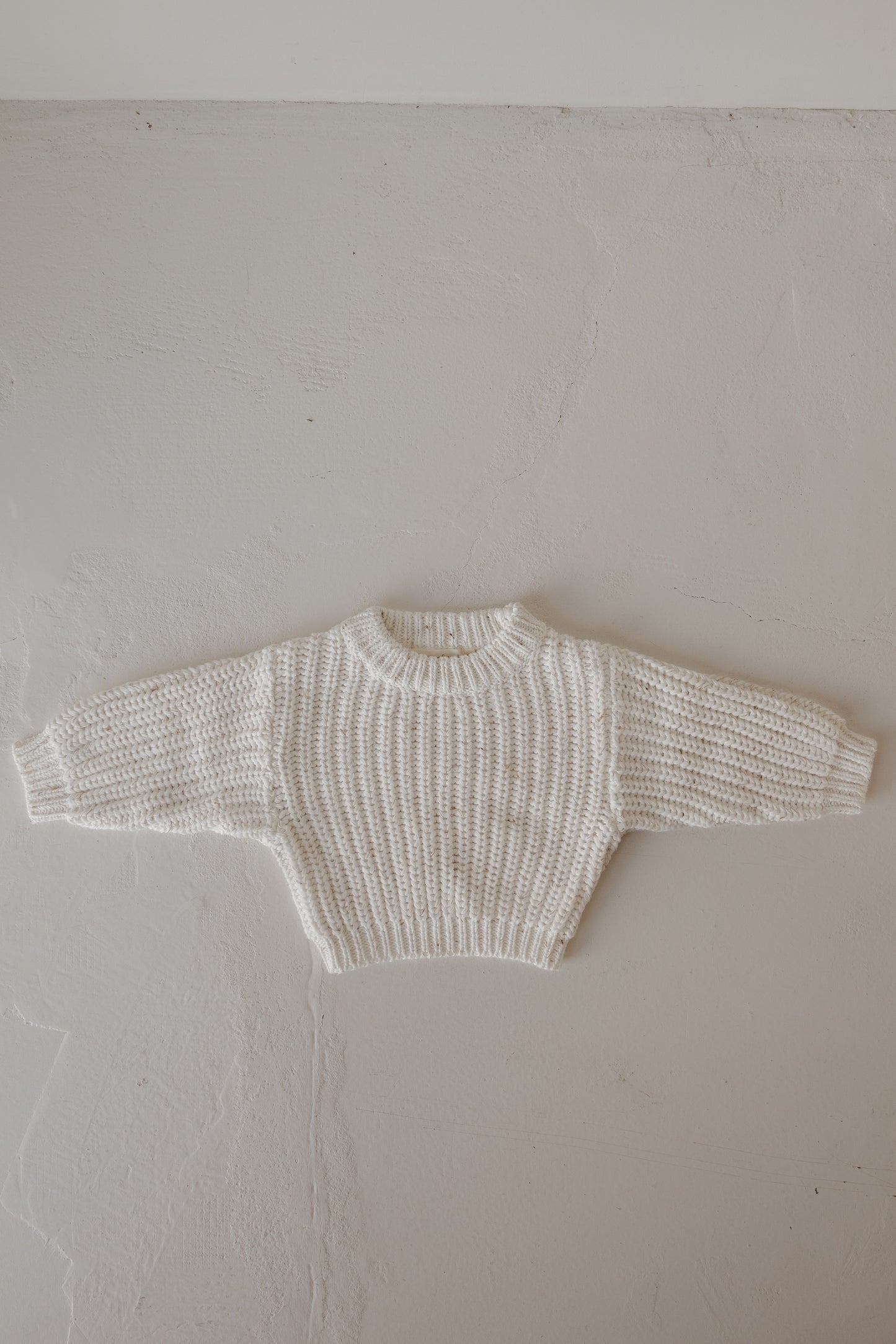 The Child Knit Sweater | Cloud Confetti by forever french baby is a cream-colored, hand-knitted sweater with long sleeves, a ribbed pattern, and a round neckline, shown laid flat on a textured light gray surface.