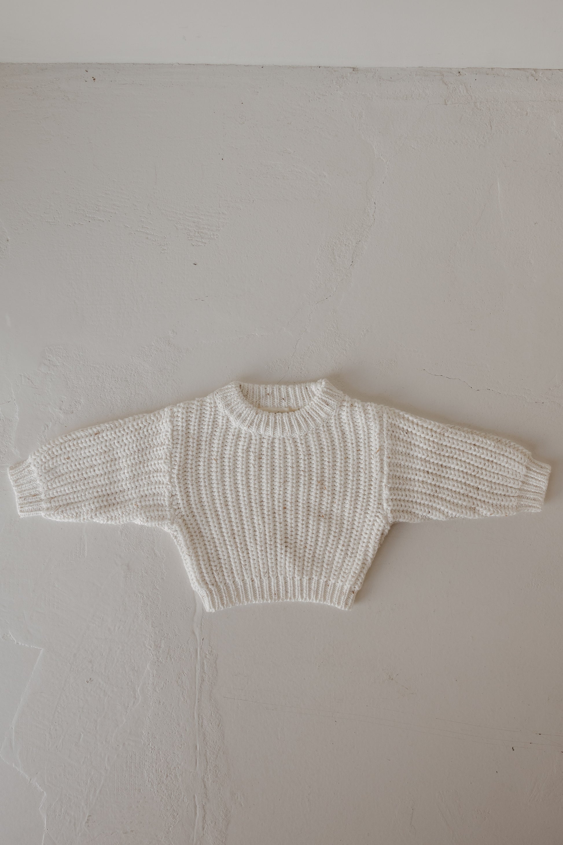 The Child Knit Sweater | Cloud Confetti by forever french baby is a cream-colored, hand-knitted sweater with long sleeves, a ribbed pattern, and a round neckline, shown laid flat on a textured light gray surface.