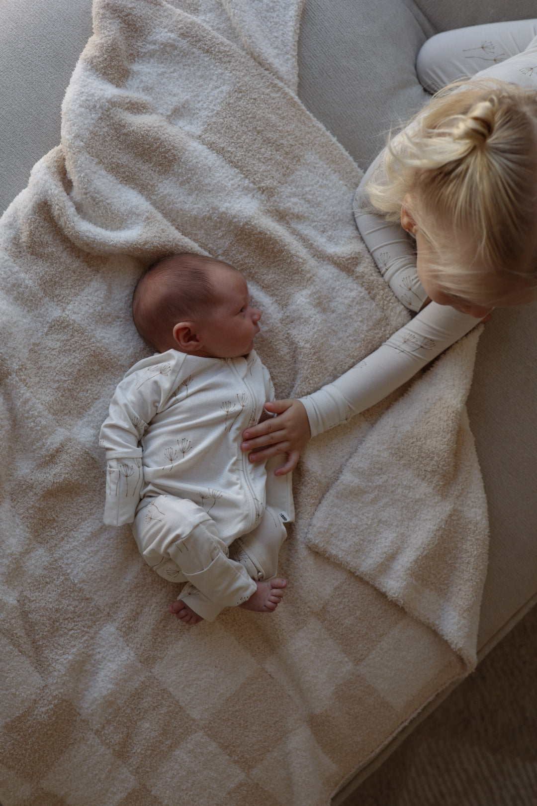 A baby in forever french baby's Bamboo Two Piece Pajamas | Dandelion rests on a beige blanket, while a blonde-haired child gently touches the baby's chest as they relax together on a cozy couch.