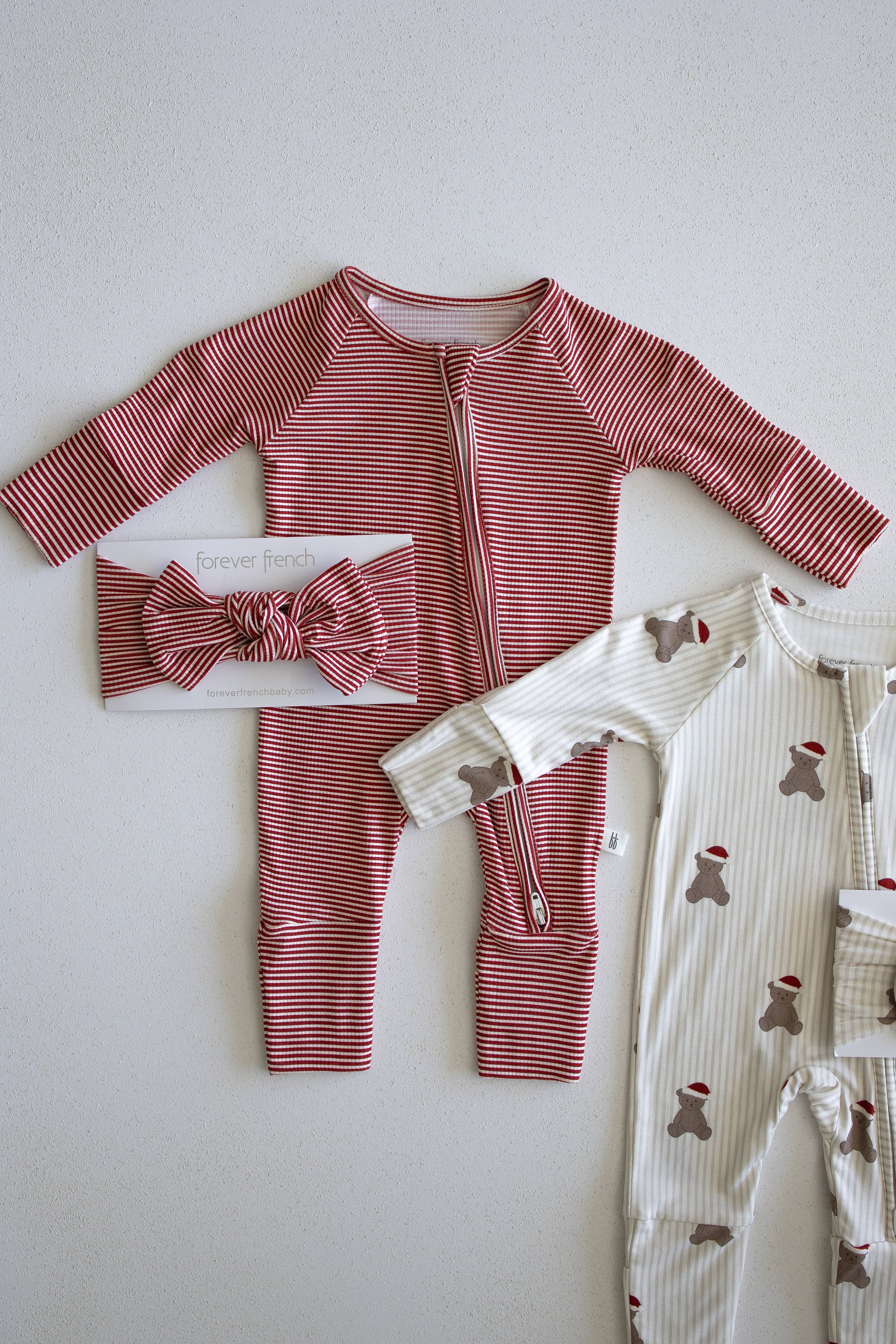 A pair of forever french baby Bamboo Zip Pajamas in Peppermint Stripe Ribbed—a red and white striped baby onesie—paired with a matching headband, is displayed alongside a cream-colored hypo-allergenic baby onesie adorned with teddy bears in red hats. Both pieces are laid flat on a light background and crafted from soft, ribbed bamboo fabric designed for gentle comfort on delicate skin.