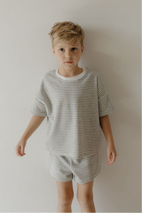 A young child with short blonde hair stands against a white wall, wearing the forever french baby Child Terry Short Set in Grey Stripe, looking to the side with a neutral expression—perfect for cozy family moments.