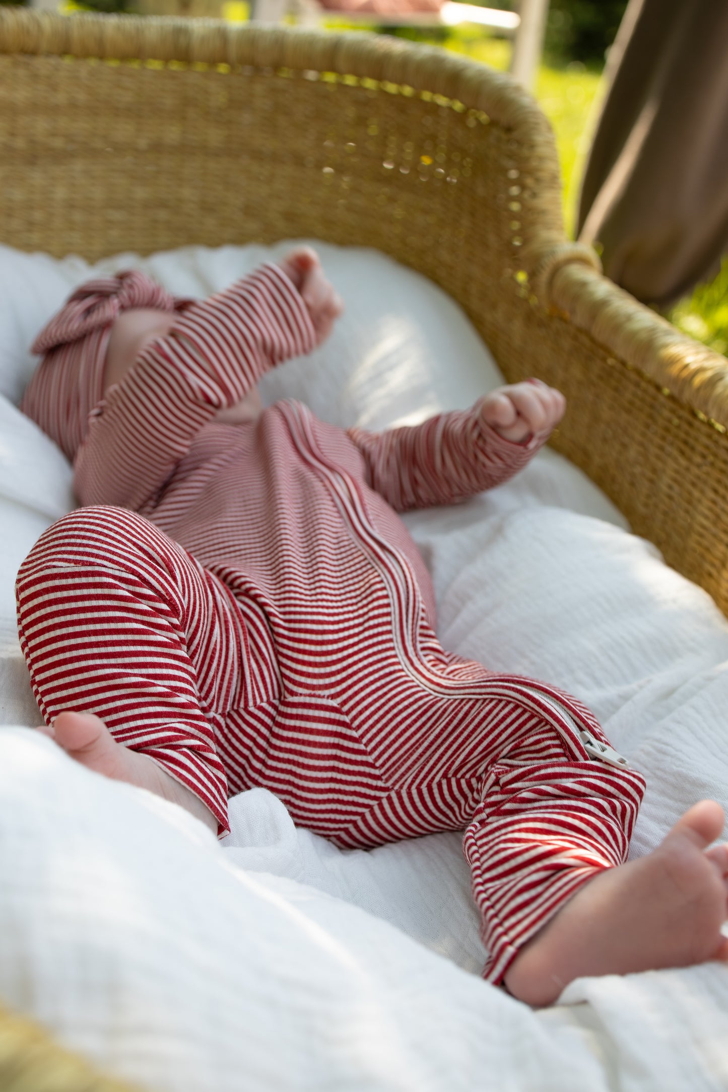 Clad in forever french baby's Bamboo Zip Pajamas | Peppermint Stripe Ribbed, a baby rests in a wicker bassinet atop a white blanket, sunlight streaming softly from the side.