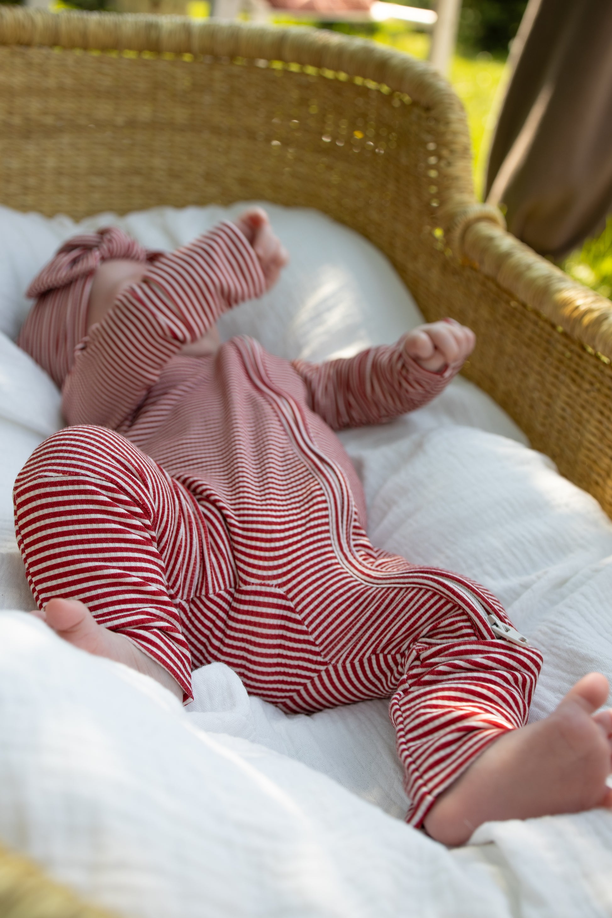 Clad in forever french baby's Bamboo Zip Pajamas | Peppermint Stripe Ribbed, a baby rests in a wicker bassinet atop a white blanket, sunlight streaming softly from the side.