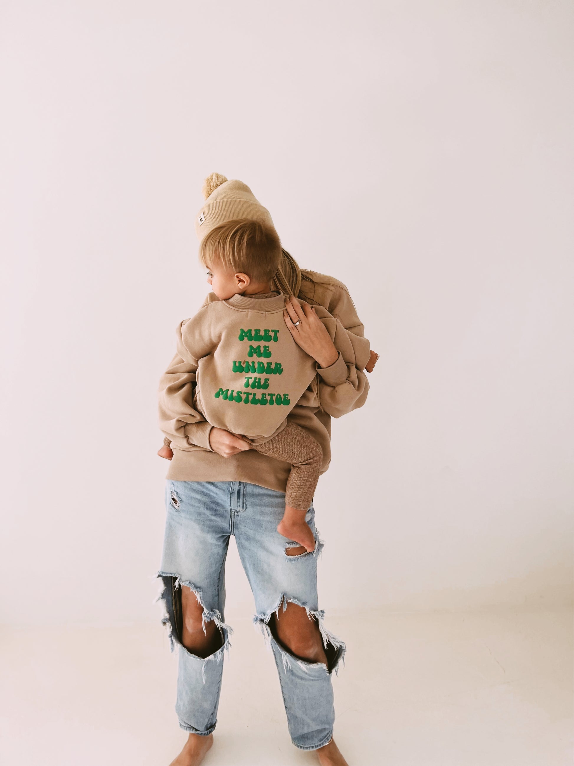 An adult, barefoot and wearing ripped jeans, holds a baby dressed in a festive sweatshirt from forever french baby that reads "Meet Me Under The Mistletoe" in green text. The adult is casually dressed in a beige beanie and sweatshirt.