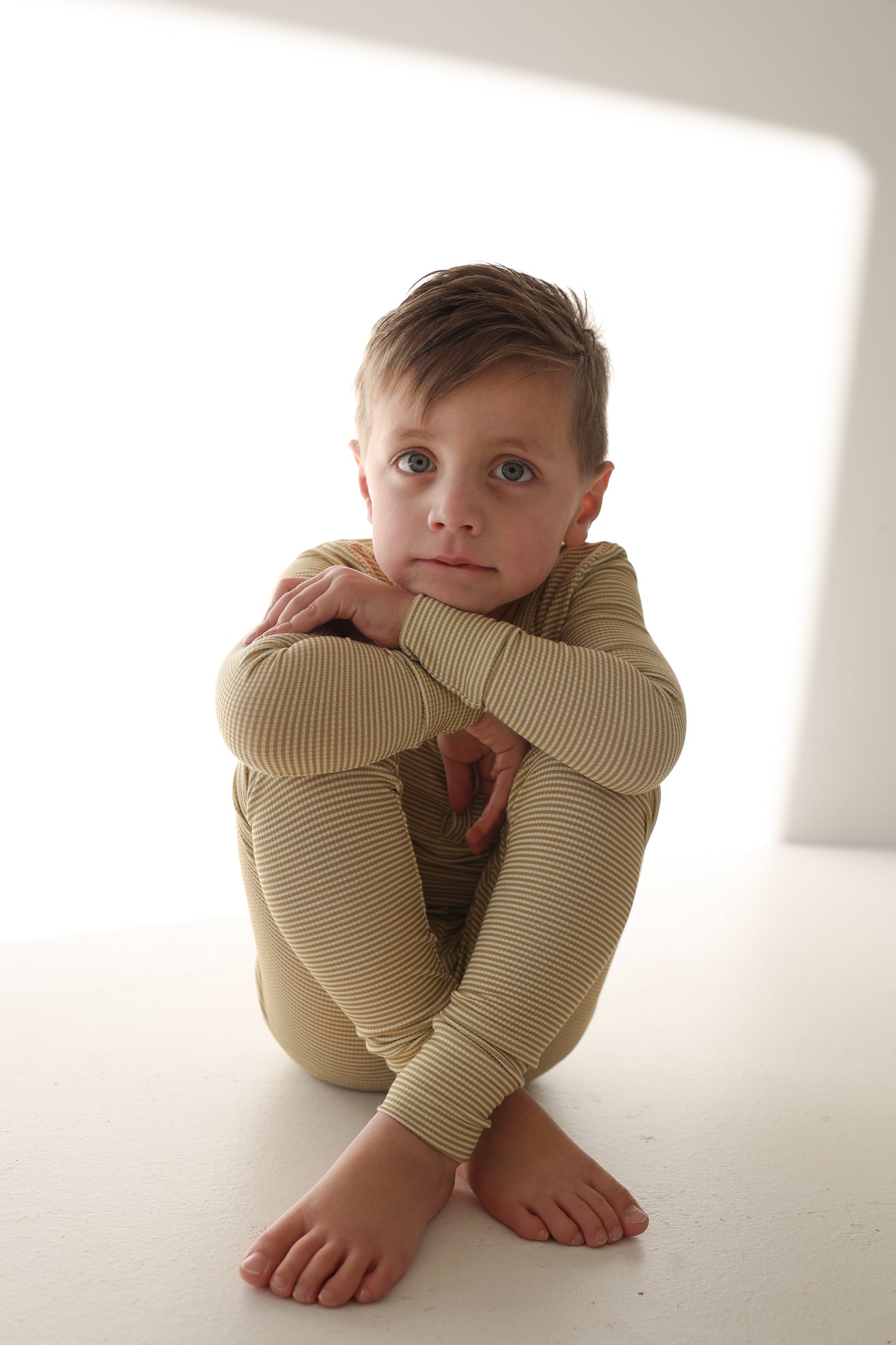 A young boy with light brown hair sits barefoot on a white surface, hugging his knees and looking at the camera. He is wearing forever french baby Bamboo Two Piece Pajamas | Willow Stripe Ribbed—beige, striped long sleeves and pants.