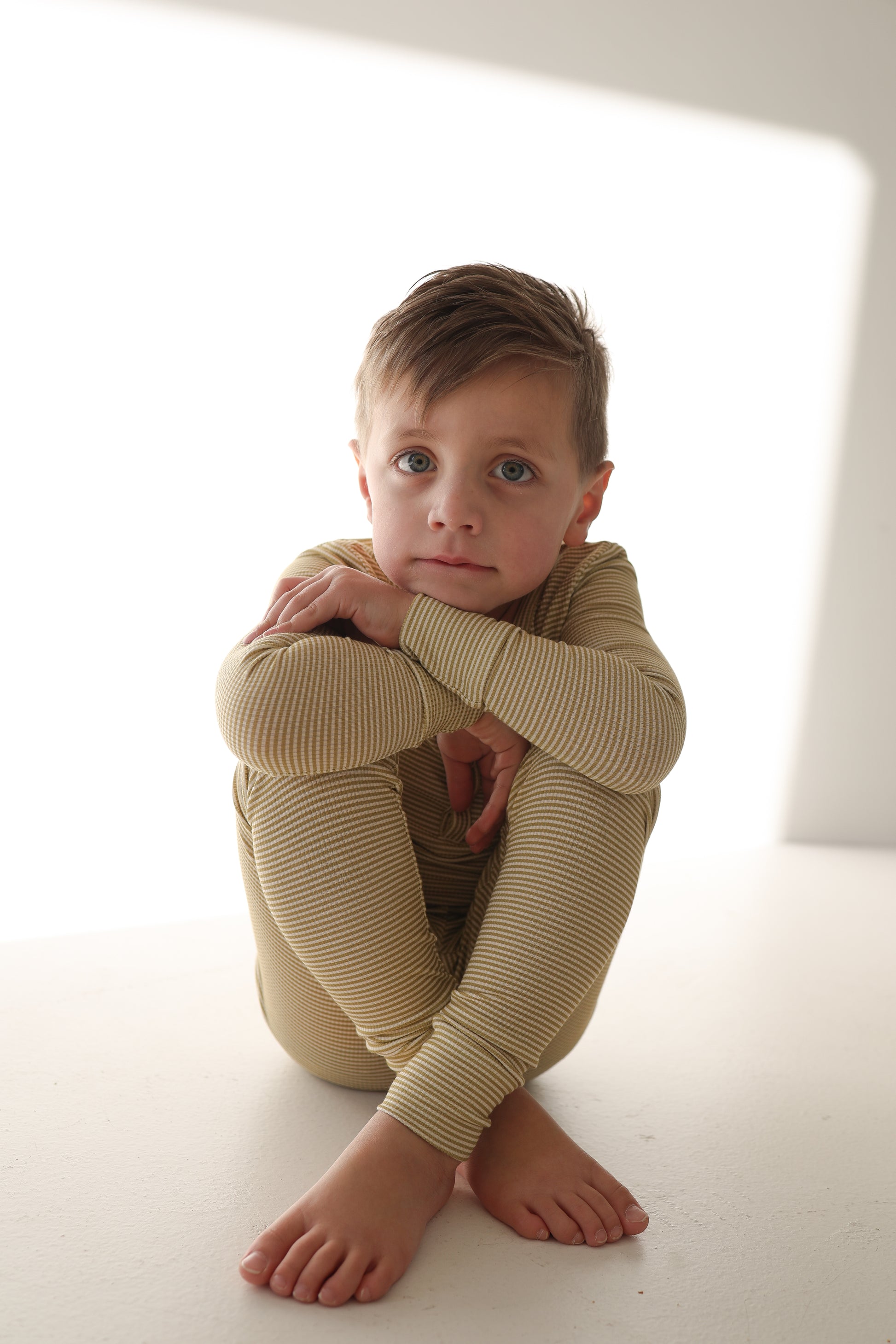 A young boy with light brown hair sits barefoot on a white surface, hugging his knees and looking at the camera. He is wearing forever french baby Bamboo Two Piece Pajamas | Willow Stripe Ribbed—beige, striped long sleeves and pants.