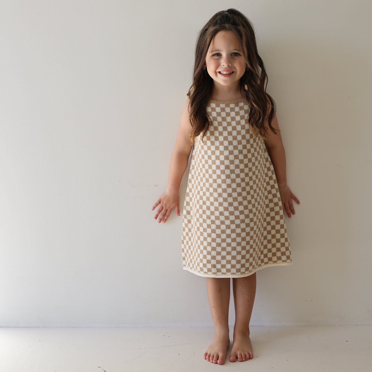 A young girl with long brown hair stands barefoot against a white wall, smiling and wearing the Knit Dress | Latte Check from forever french baby’s cotton kids clothing collection.