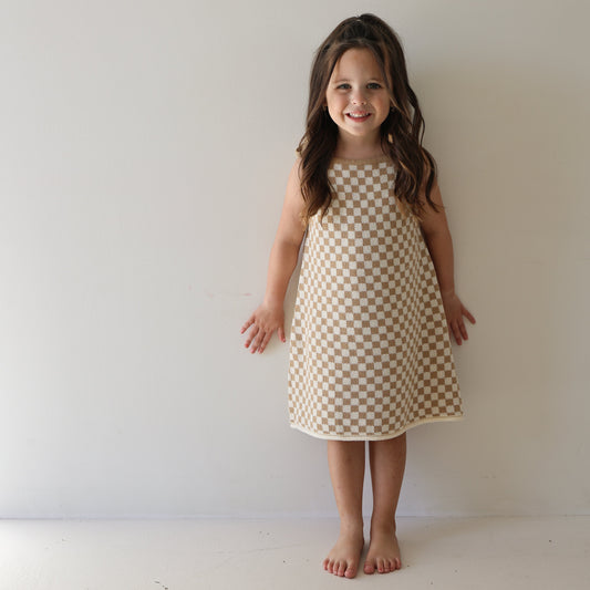 A young girl with long brown hair stands barefoot against a white wall, smiling and wearing the Knit Dress | Latte Check from forever french baby’s cotton kids clothing collection.