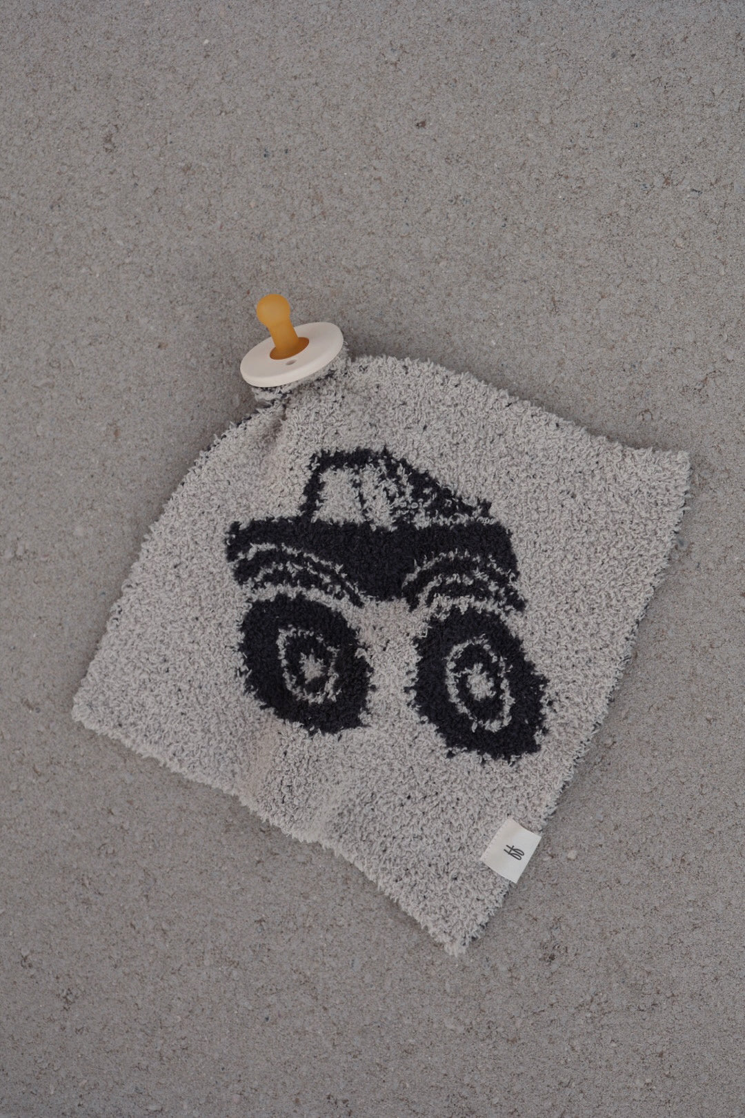 The forever french baby Lovey | Monster Truck, a gray plush microfiber lovey featuring a black monster truck cartoon and an attached beige pacifier, rests on a speckled light gray surface.