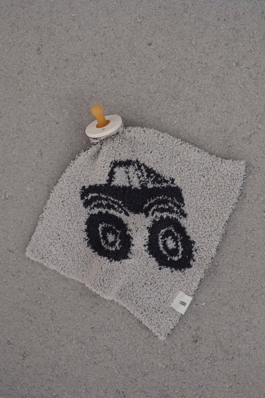 The forever french baby Lovey | Monster Truck, a gray plush microfiber lovey featuring a black monster truck cartoon and an attached beige pacifier, rests on a speckled light gray surface.