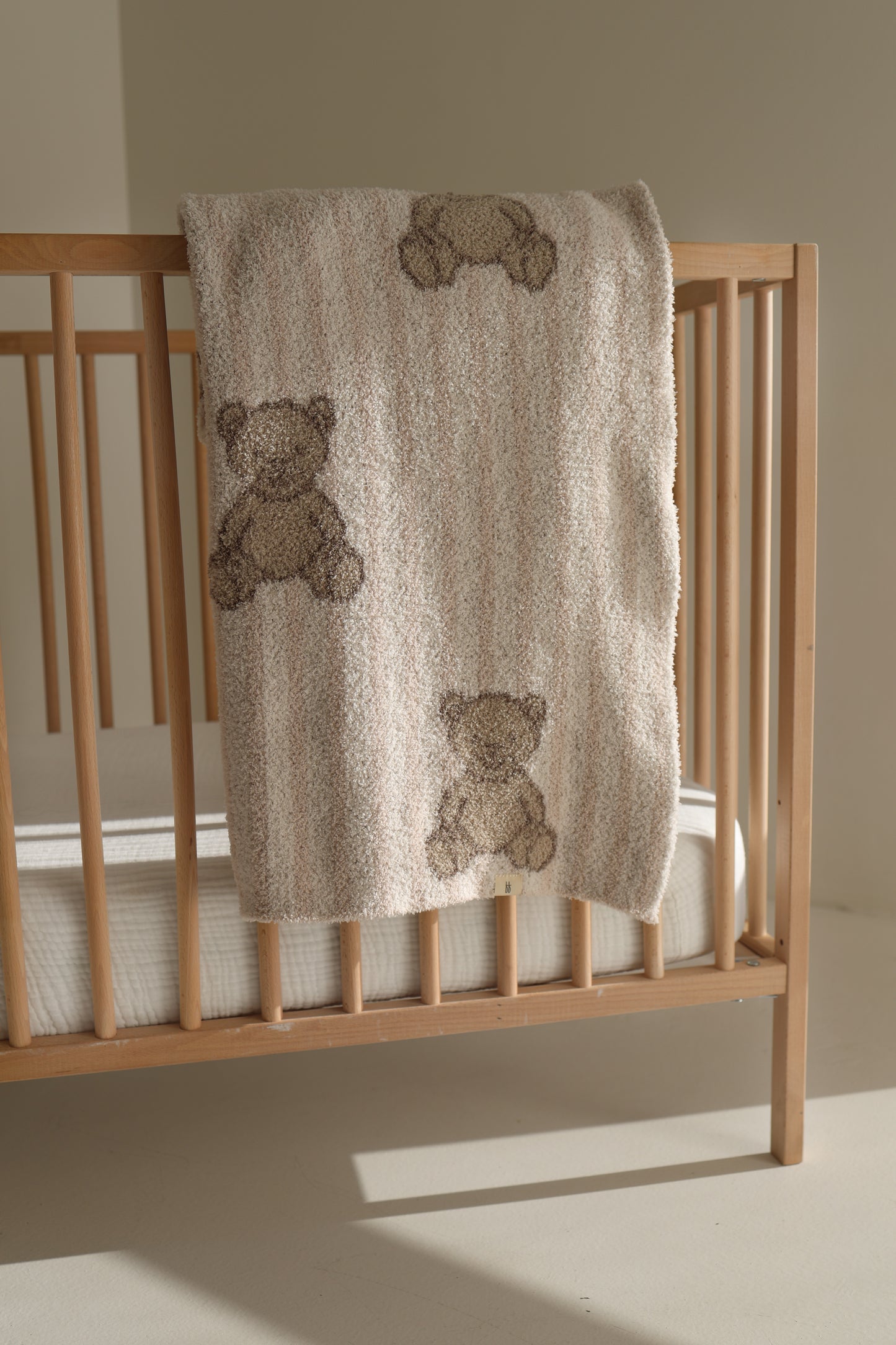 A wooden crib with a white mattress features the forever french baby Plush Blanket | Teddy, an Oeko-Tex certified beige blanket with brown teddy bear designs, draped over the rail and bathed in natural sunlight.