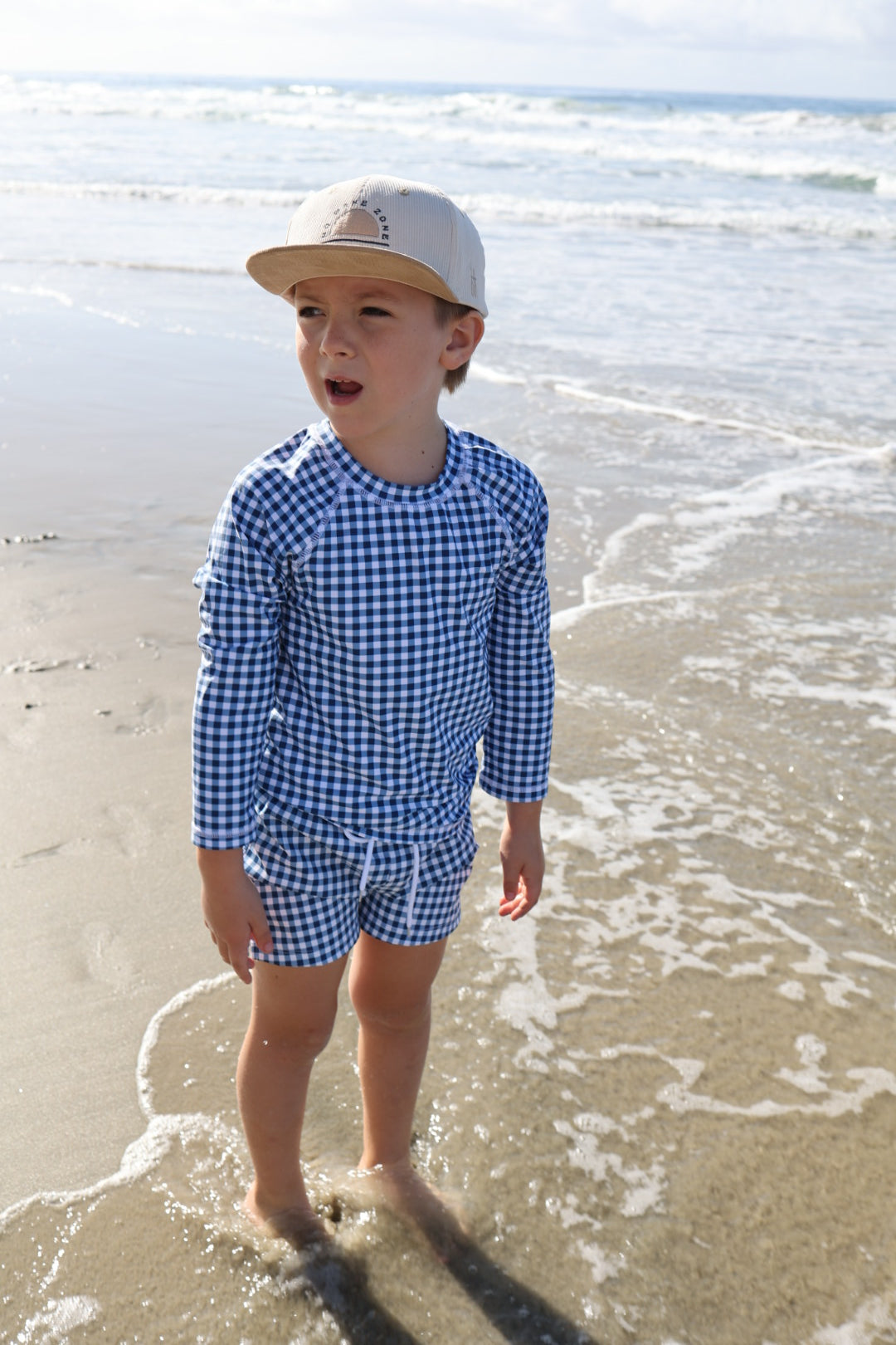 A young boy wearing forever french baby’s Child Board Short in Blue Gingham stands at the edge of the ocean, waves softly touching his feet on a sunny beach.