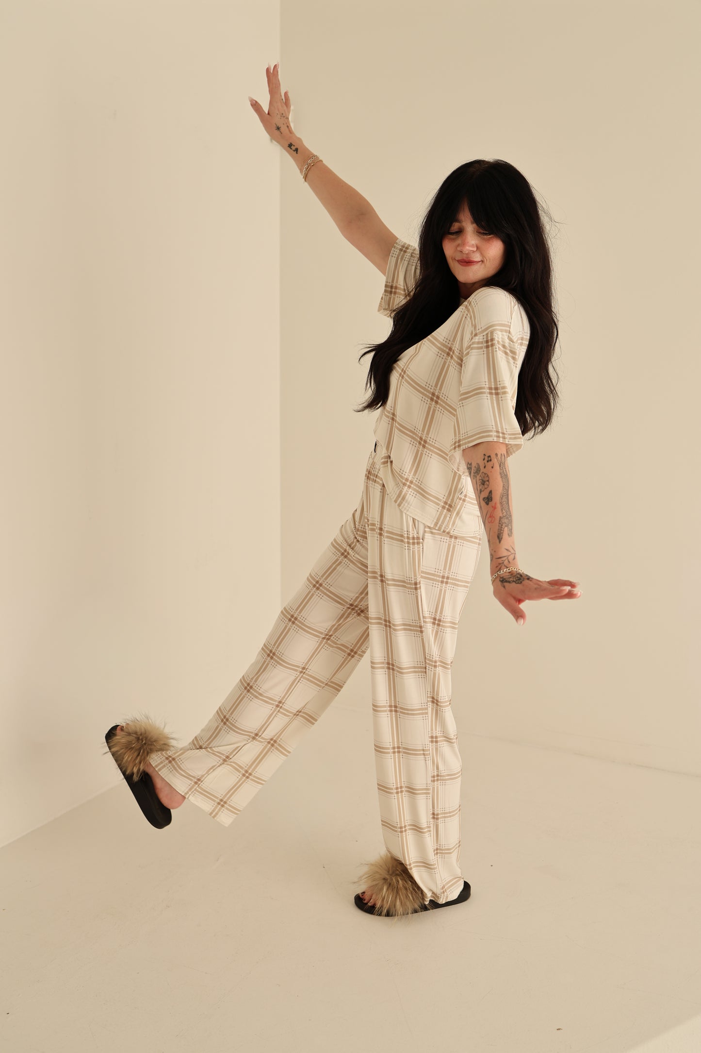 A woman with long dark hair poses playfully in a minimal beige room, wearing the Women's Boxy Tee Lounge Set | Ellis Plaid by forever french baby—smiling as she showcases this cozy, hypo-allergenic style.