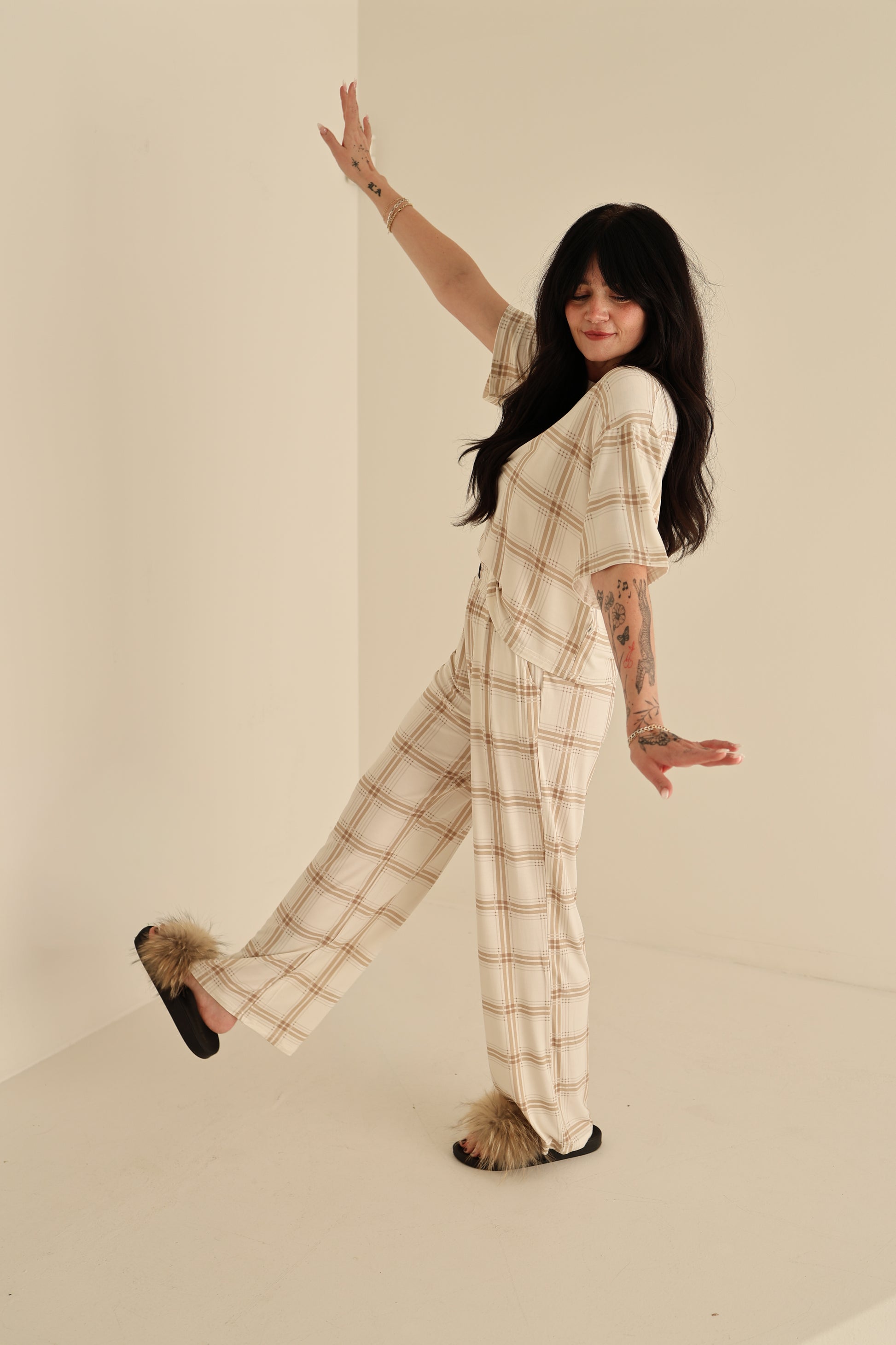 A woman with long dark hair poses playfully in a minimal beige room, wearing the Women's Boxy Tee Lounge Set | Ellis Plaid by forever french baby—smiling as she showcases this cozy, hypo-allergenic style.