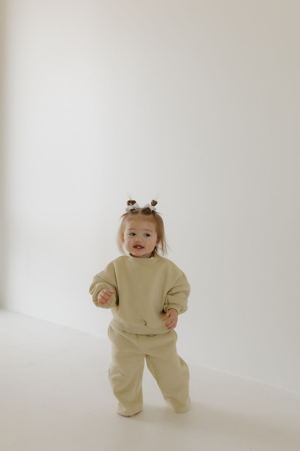 A toddler wearing a forever french baby Child Sweat Set | All the Good Things stands indoors on a white floor against a light-colored wall. The toddler has small pigtails and is looking forward with a cheerful expression.