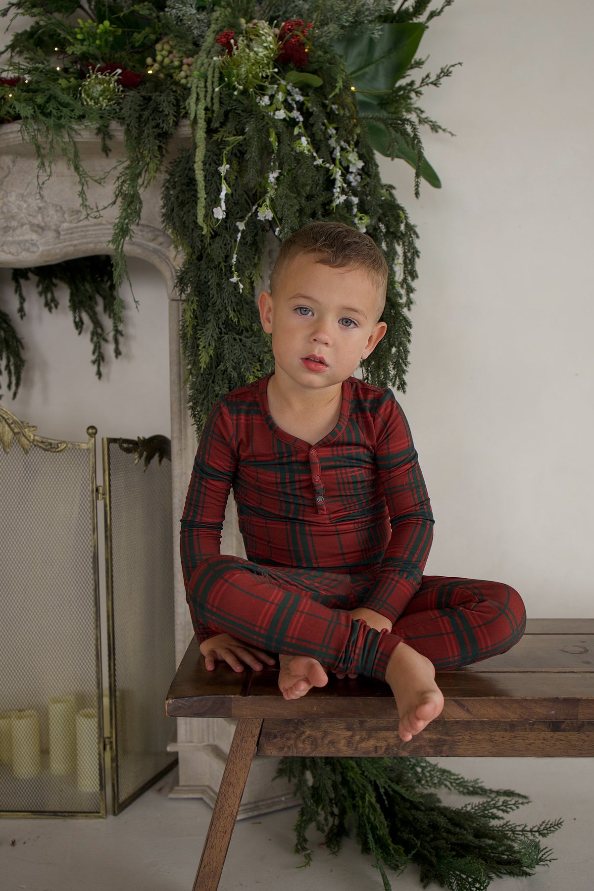A young boy sits on a wooden bench wearing a long-sleeved, Christmas plaid Bamboo Two Piece Pajamas set by forever french baby, featuring buttons at the collar.
