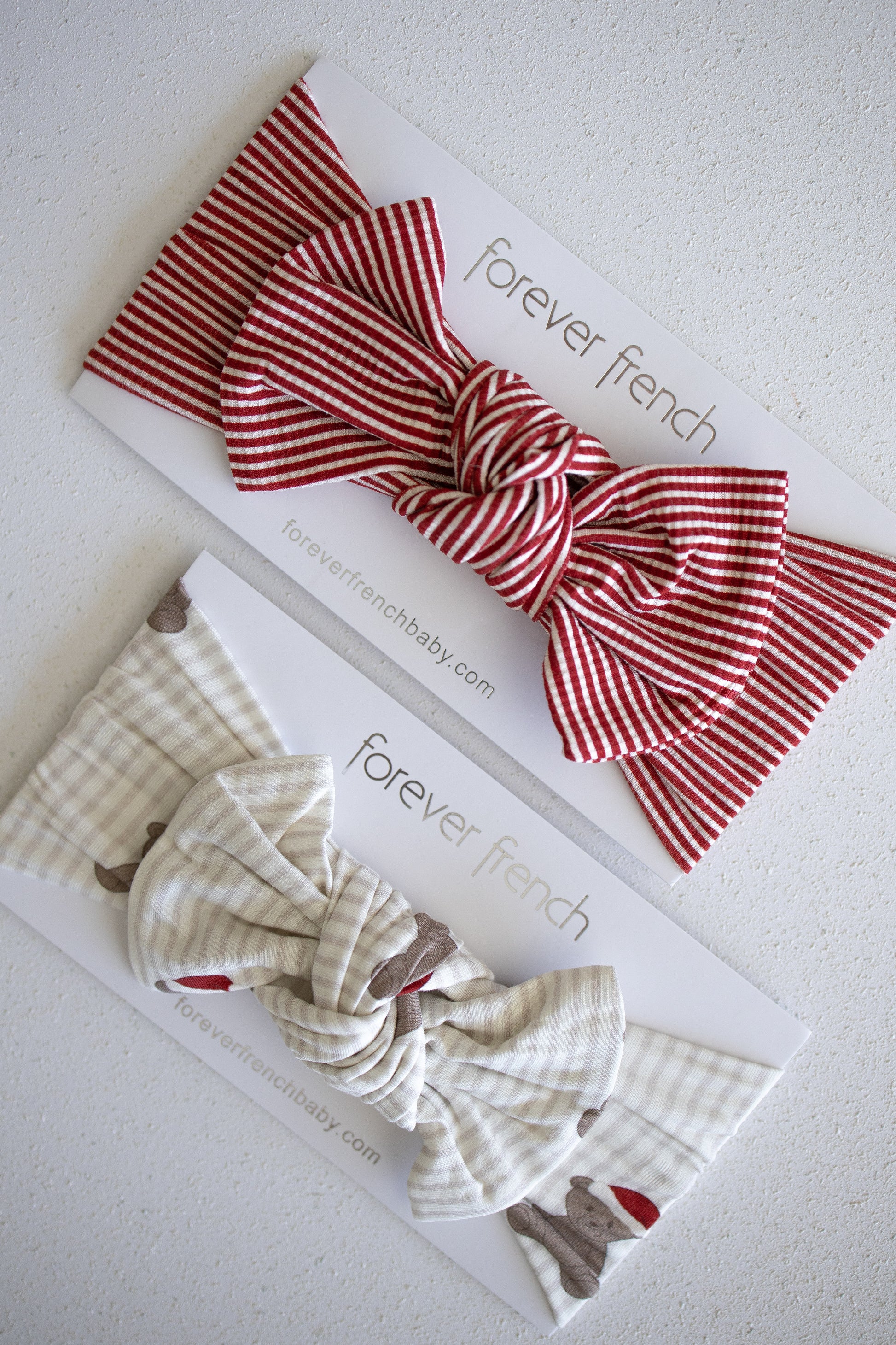 Two forever french baby bamboo head wraps with large bows are displayed on branded cards—a Peppermint Stripe Ribbed style in red and white, and a cream-colored one with a subtle pattern—shown on a light surface.