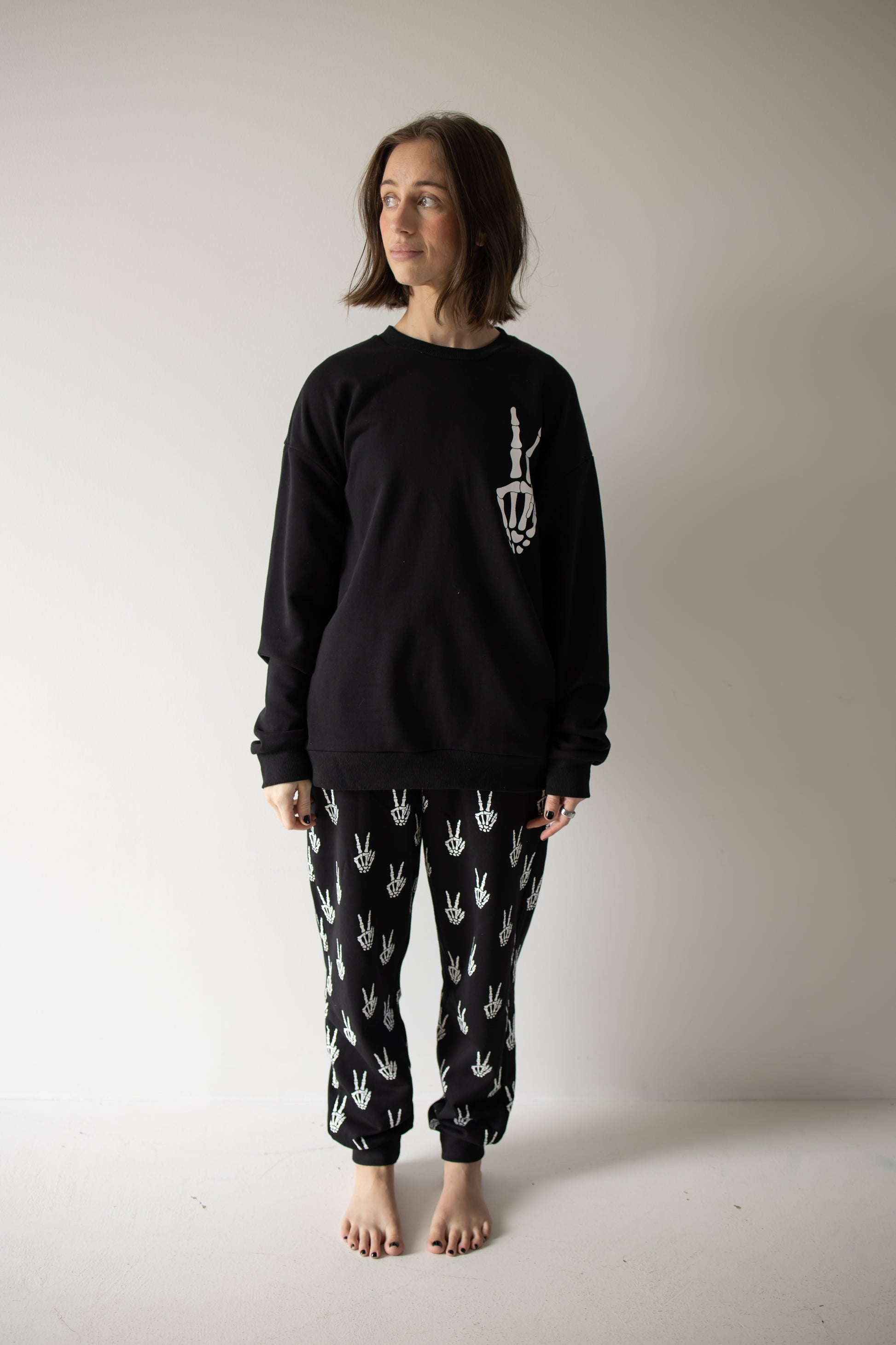 Barefoot against a white wall, a woman with shoulder-length brown hair models the Women’s Sweat Set | Midnight Skelly from forever french baby—an effortlessly cool black set featuring striking white handprint designs, perfect for spooky season. She gazes slightly to the left with a subtle smile, adding modern edge to your go-to pumpkin patch outfit.
