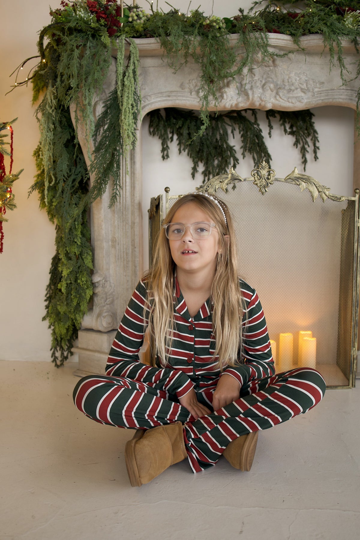 A girl wearing a Girl’s Bamboo Button Up Lounge Set | Holiday Stripe and tan boots sitting cross-legged in front of a fireplace.